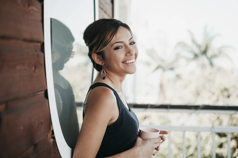 Una mujer sonriendo junto a una ventana con luz de mañana, con una taza en la mano.