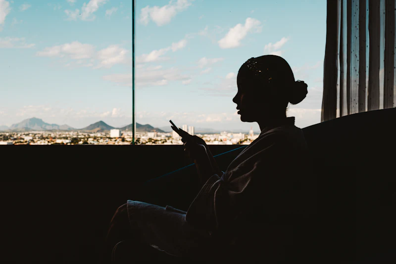 Silueta de una mujer mirando el móvil junto a una ventana, con el cielo azul detrás.